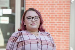 Eine Frau mit pinkem Haar und Brille steht vor einer Ziegelmauer.