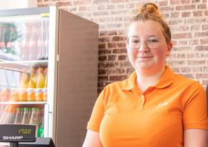 Eine Frau in einem orangefarbenen Hemd steht vor einem Kühlschrank mit Getränken.